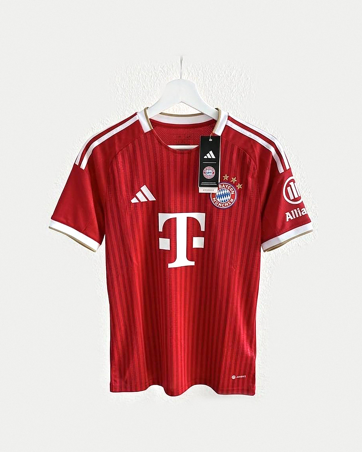 Camiseta de fútbol roja con rayas verticales a tono, franjas blancas y ribete dorado en los hombros, cuello blanco y escudo bordado con tres estrellas, colgada en una percha sobre pared clara