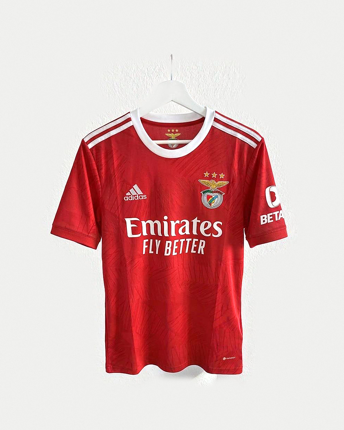 Camiseta de fútbol roja con textura a tono, cuello redondo blanco, franjas blancas en los hombros y escudo bordado con tres estrellas encima, colgada en una percha sobre pared clara