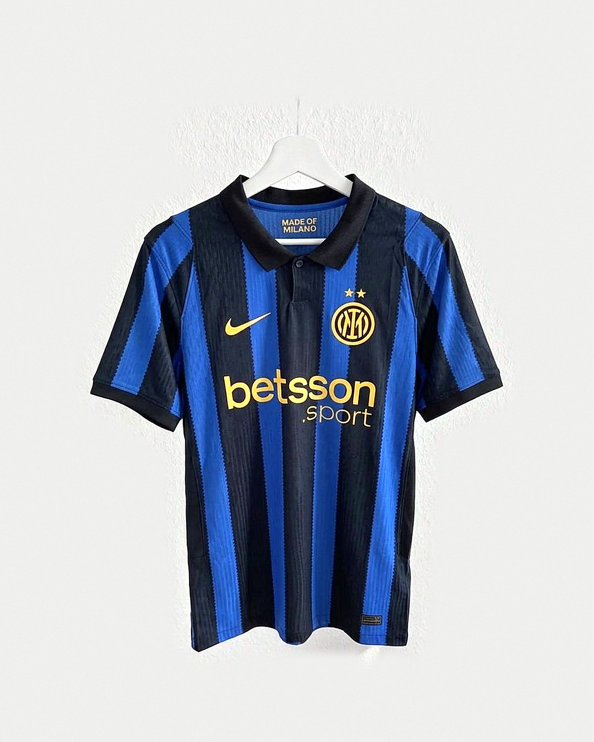 Camiseta de fútbol a rayas verticales azules y negras con cuello polo negro y escudo bordado en dorado con dos estrellas, colgada en una percha sobre pared clara