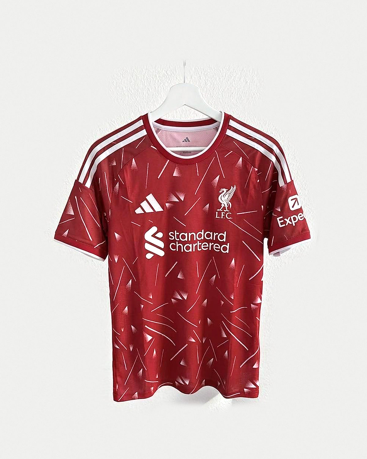 Camiseta de fútbol roja con estampado geométrico rosa claro, franjas blancas en los hombros y escudo bordado con un ave, colgada en una percha sobre pared clara