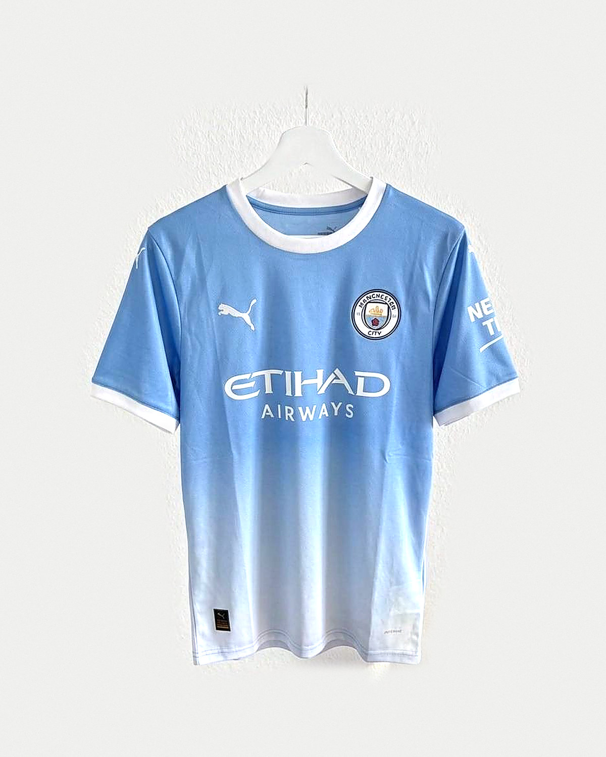 Camiseta de fútbol azul celeste degradada a blanco en el bajo, con cuello y puños blancos y escudo circular bordado, colgada en una percha sobre pared clara