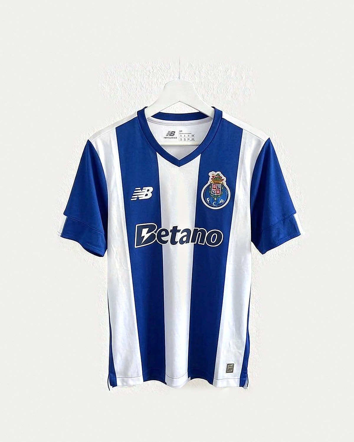 Camiseta de fútbol a franjas verticales anchas azules y blancas, con cuello de pico azul, mangas azules de puño blanco y escudo bordado en el pecho, colgada en una percha sobre pared clara