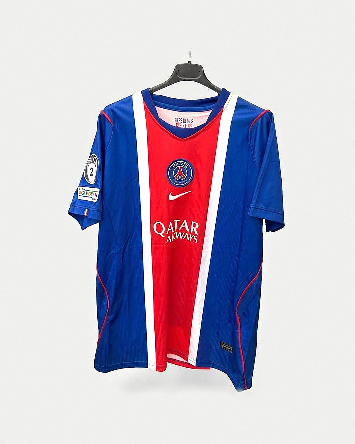 Camiseta de fútbol azul con una franja central roja ancha ribeteada en blanco, cuello de pico con ribete blanco y rojo, escudo bordado y letras blancas grandes en el pecho y un parche circular en la manga izquierda, colgada en una percha oscura sobre fondo claro y liso