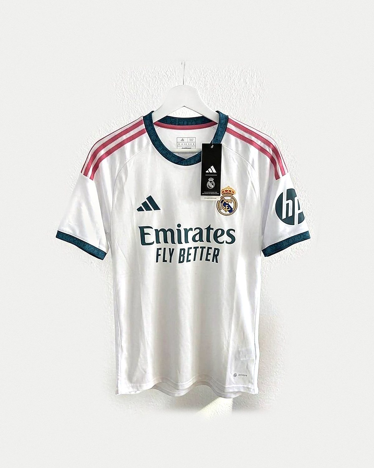 Camiseta de fútbol blanca con cuello y puños verde azulado oscuro, franjas rosas en los hombros y escudo bordado, colgada en una percha sobre pared clara