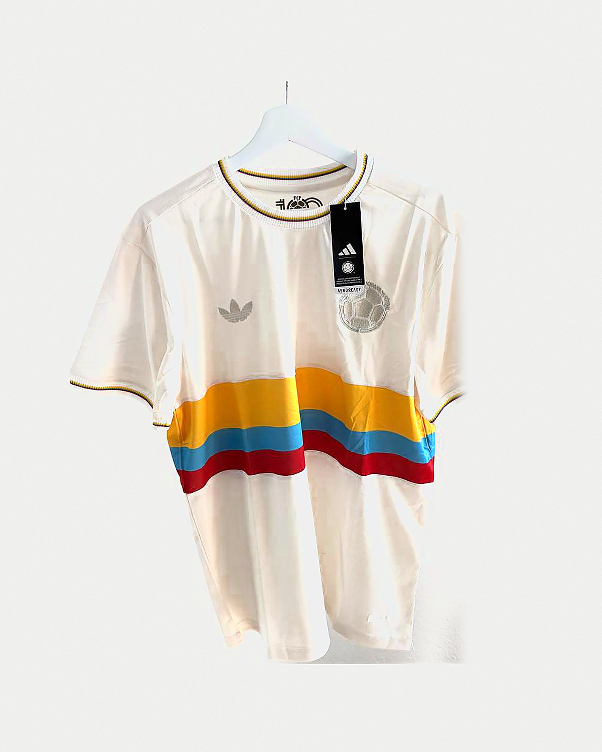 Camiseta de fútbol color crema con una banda horizontal ancha en amarillo, azul claro y rojo cruzando el pecho, escudo a tono con un balón y cuello con ribete amarillo, colgada en una percha sobre pared clara