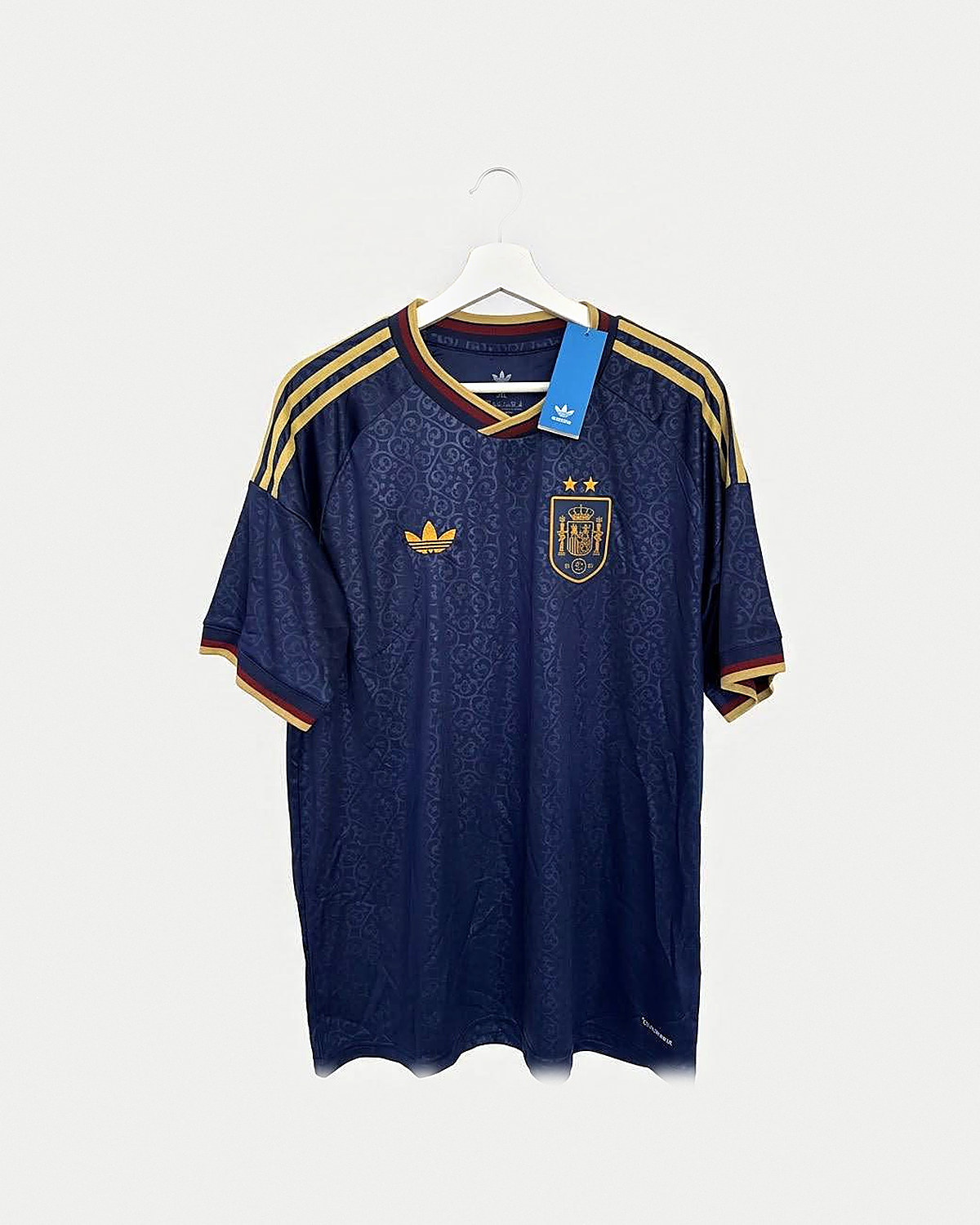 Camiseta de fútbol azul marino con filigrana a tono, franjas doradas en los hombros, cuello de pico y puños en granate y dorado y escudo dorado con dos estrellas, colgada en una percha sobre pared clara