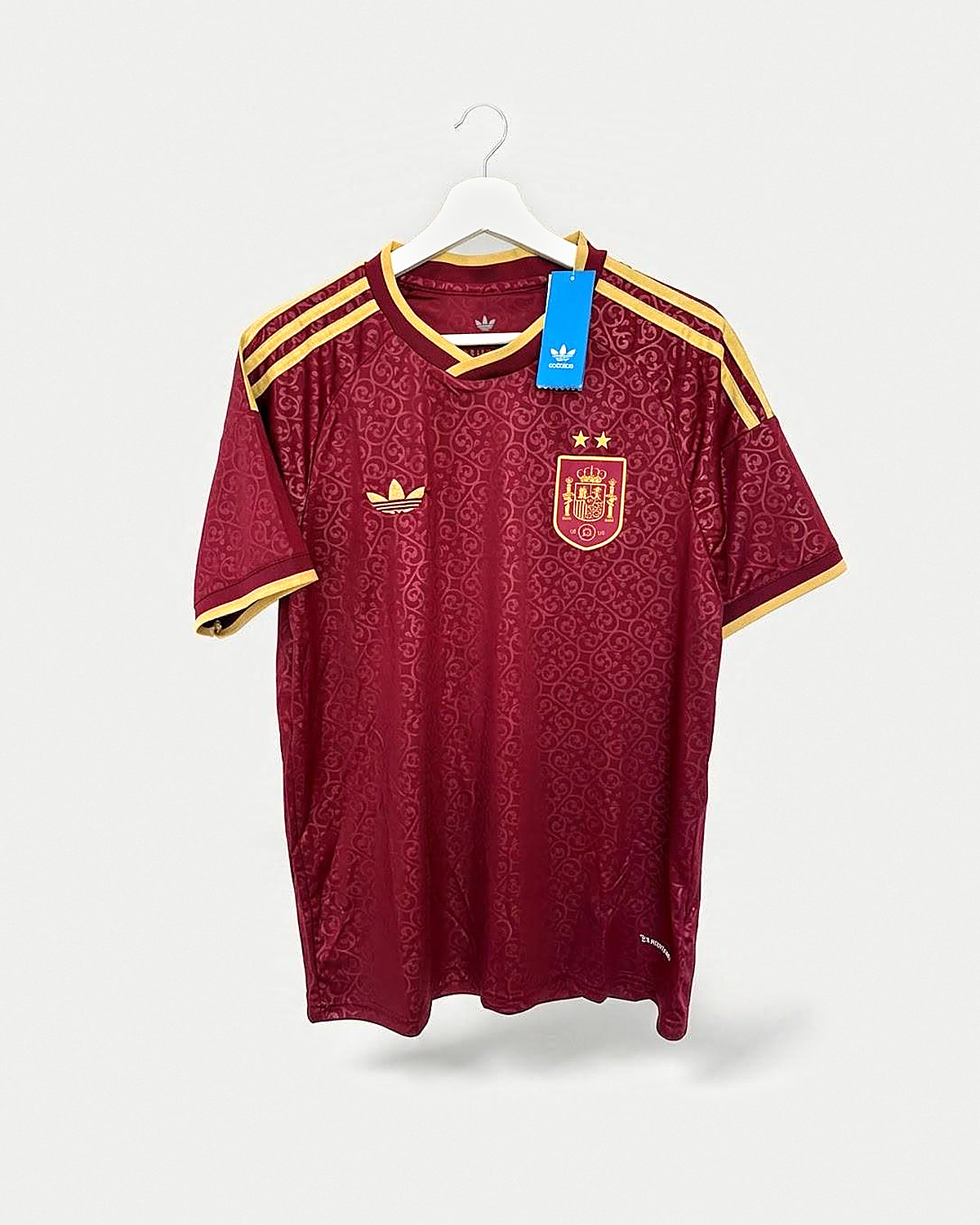 Camiseta de fútbol granate con filigrana a tono, franjas doradas en los hombros, cuello de pico y puños con ribete dorado y escudo dorado con dos estrellas, colgada en una percha sobre pared clara