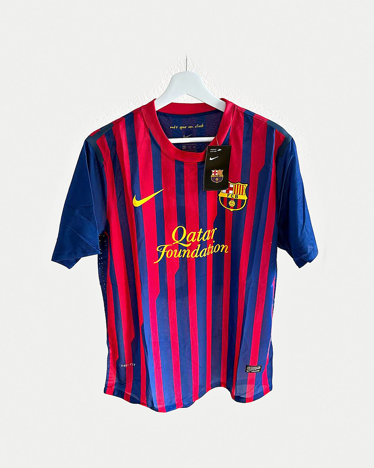 Camiseta de fútbol a rayas verticales finas azules y granates con mangas azules, cuello rojo y escudo bordado, colgada en una percha sobre pared clara