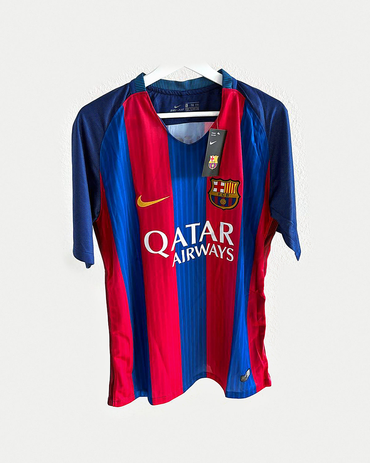 Camiseta de fútbol a franjas verticales anchas azules y granates con mangas azules y escudo bordado, colgada en una percha sobre pared clara