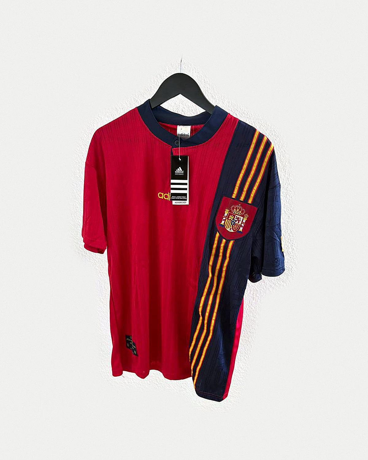 Camiseta de fútbol roja con panel diagonal azul marino y pespuntes dorados en el costado derecho y escudo bordado, colgada en una percha sobre pared clara