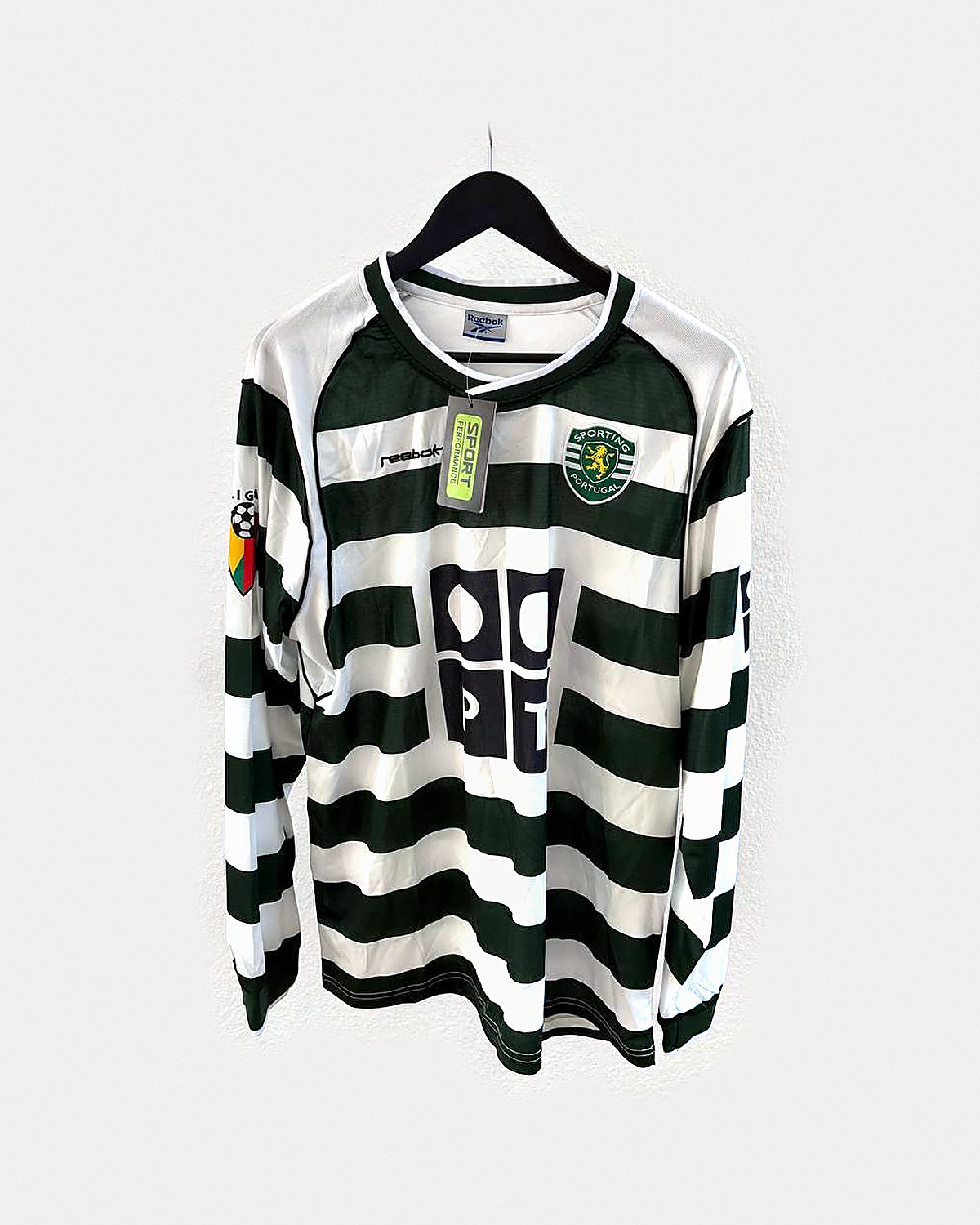 Camiseta de fútbol de manga larga a aros horizontales verde oscuro y blanco, con cuello verde y escudo bordado, colgada en una percha sobre pared clara
