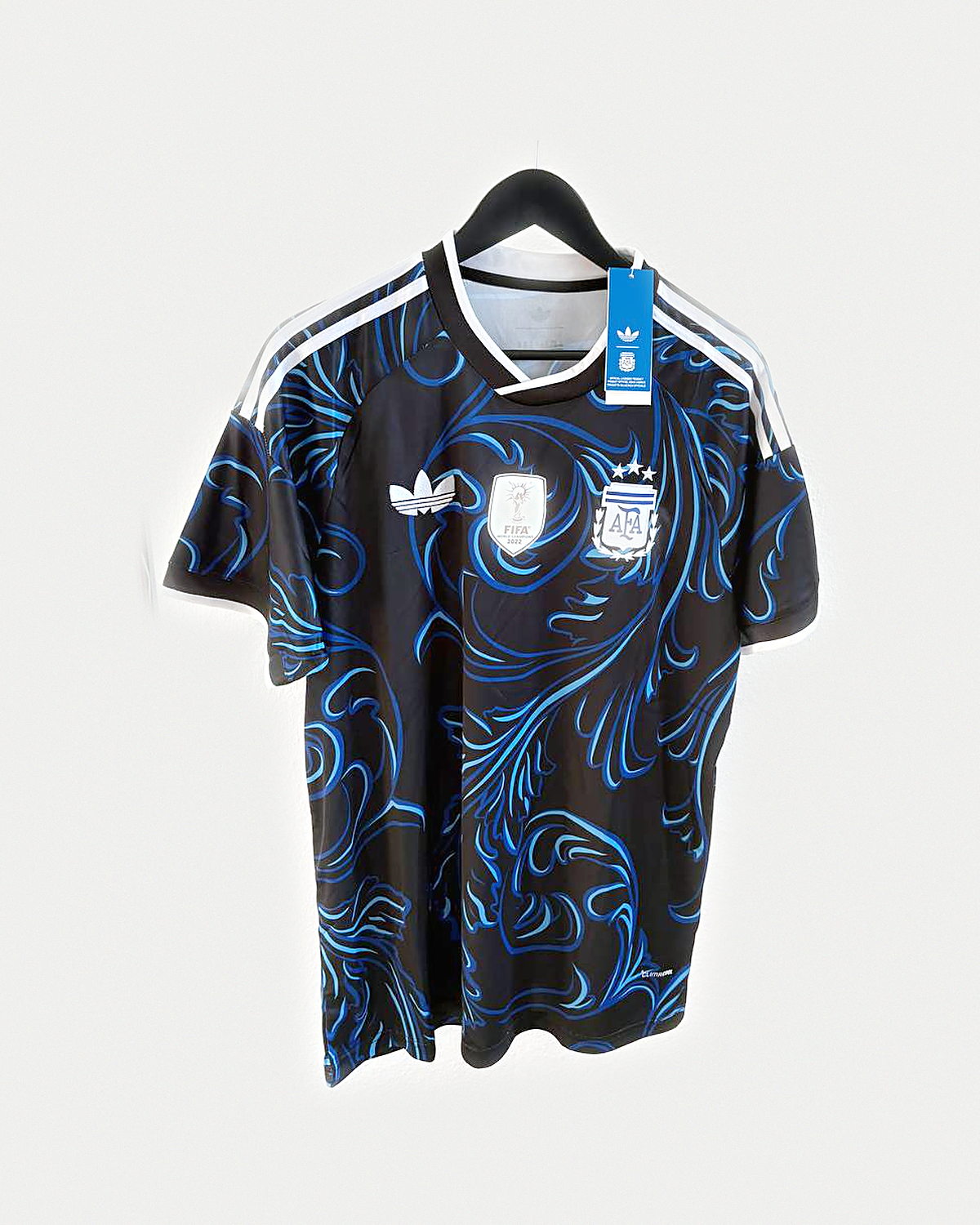 Camiseta de fútbol negra con filigrana azul y franjas blancas en los hombros, colgada en una percha sobre pared clara