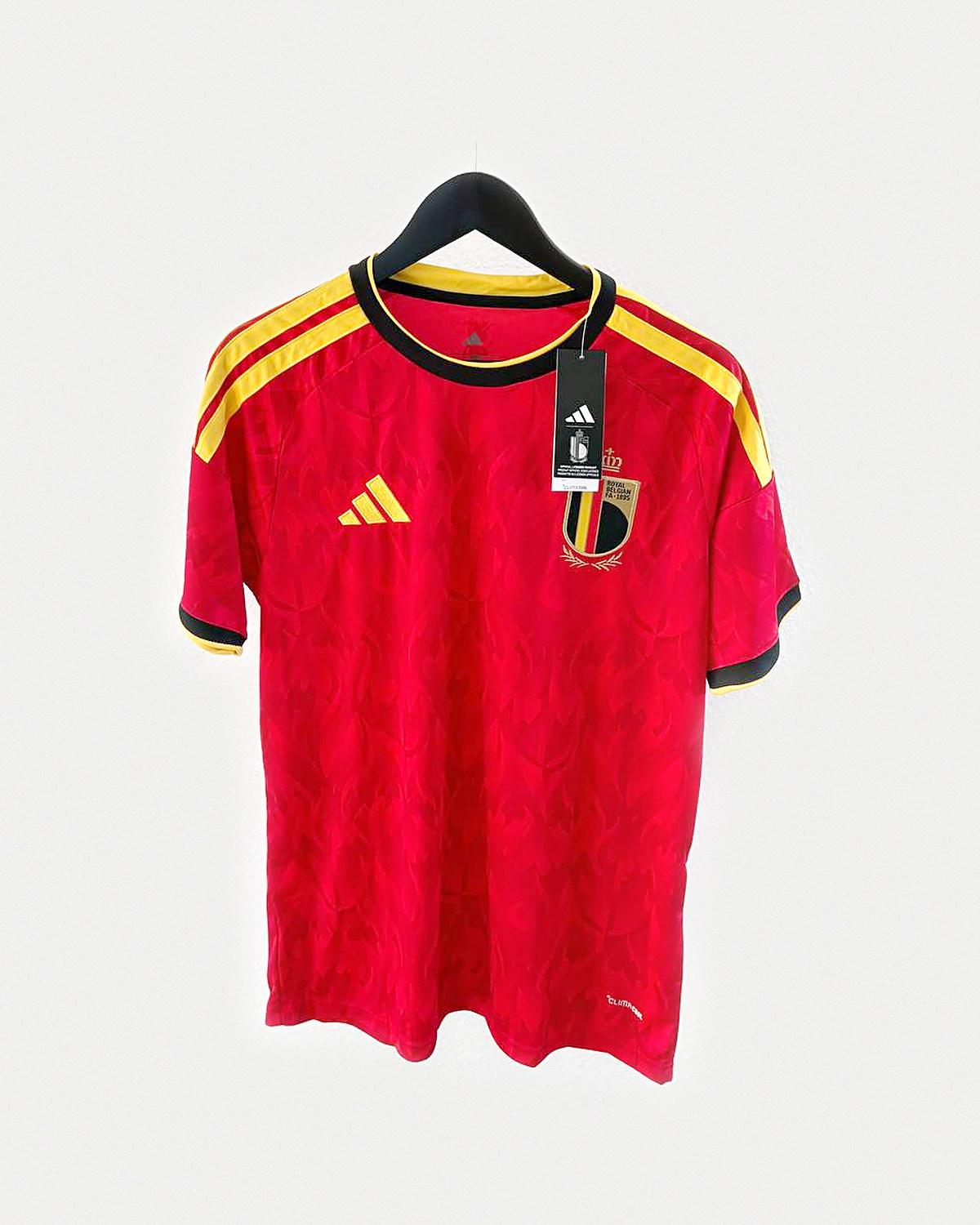 Camiseta de fútbol roja con franjas amarillas y negras en los hombros y escudo bordado, colgada en una percha sobre pared clara
