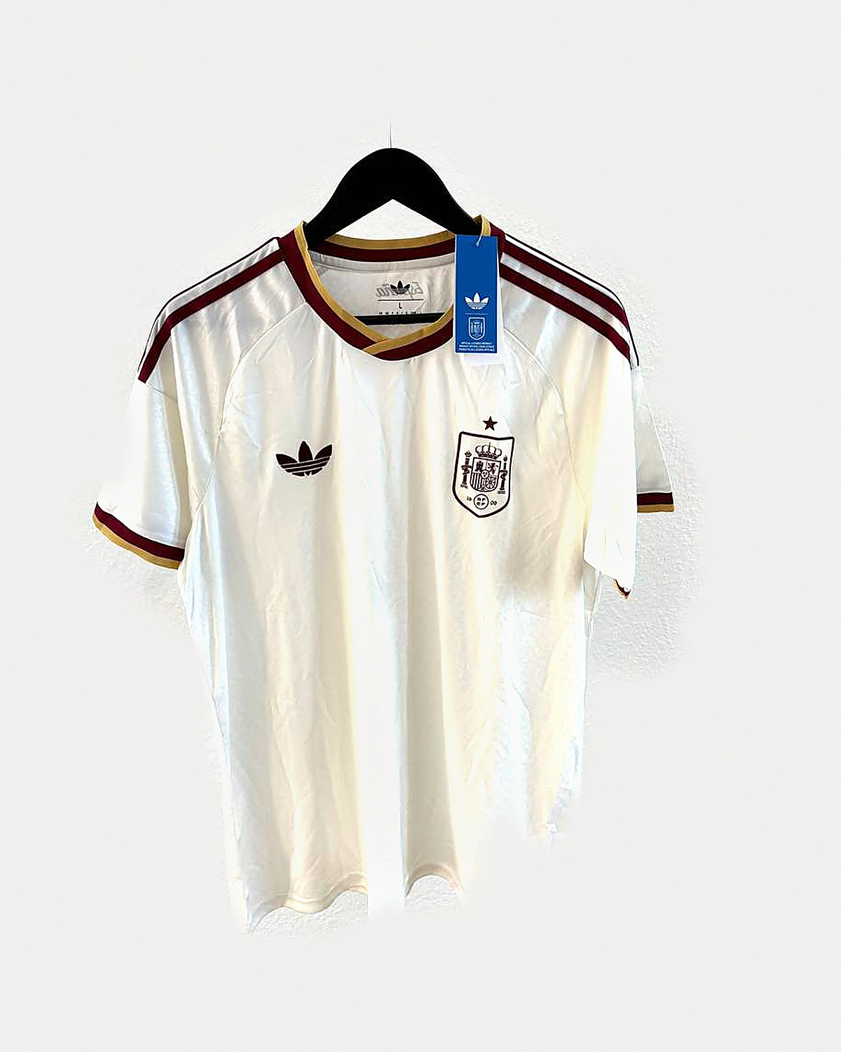 Camiseta de fútbol color crema con franjas granates y doradas en los hombros y escudo con una estrella, colgada en una percha sobre pared clara
