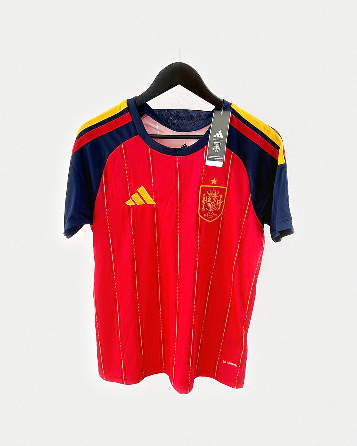 Camiseta de fútbol roja con mangas azul marino y escudo dorado con una estrella, colgada en una percha sobre pared clara