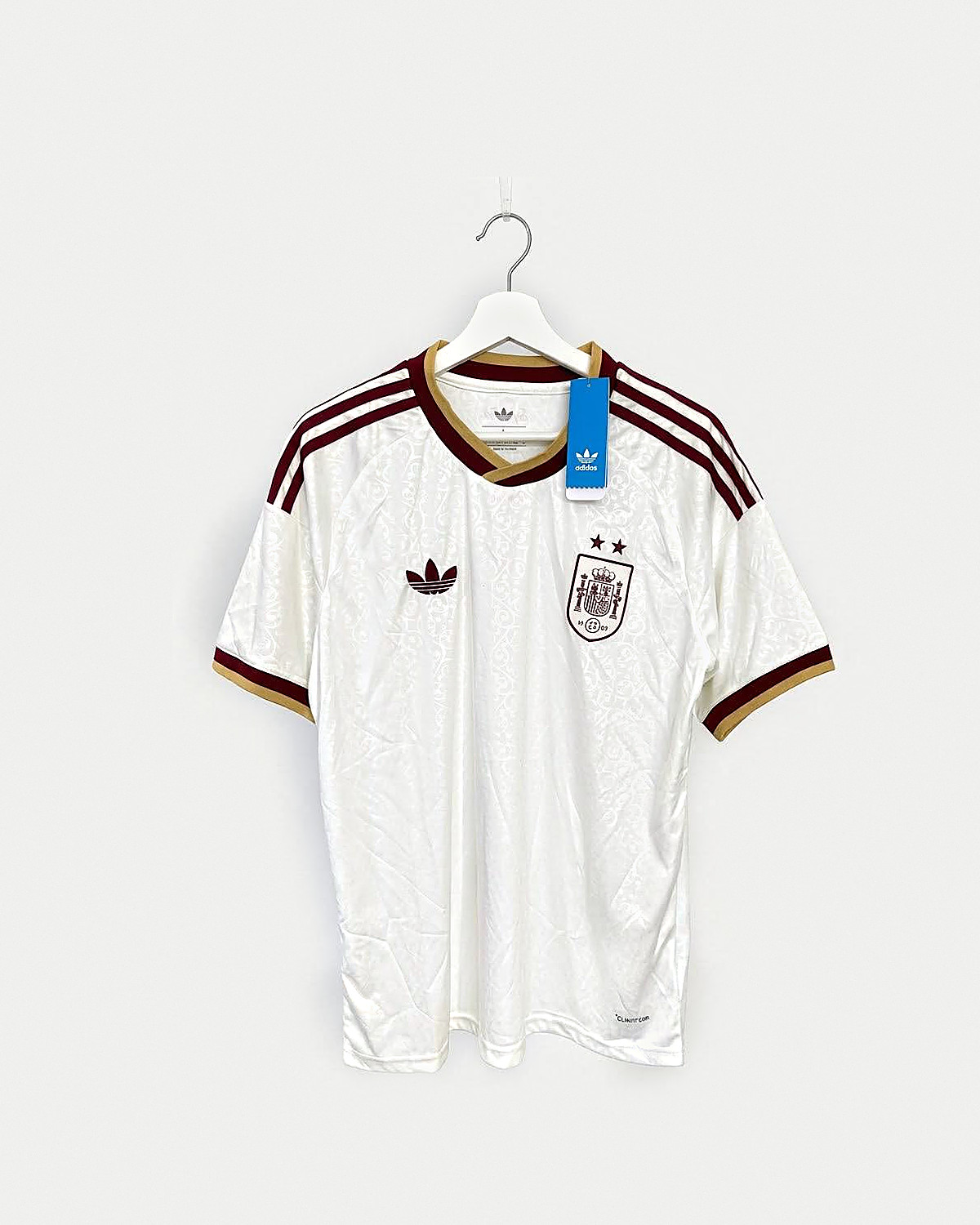 Camiseta de fútbol color hueso con filigrana a tono, franjas granates en los hombros, cuello de pico y puños en granate y dorado y escudo granate con dos estrellas, colgada en una percha sobre pared clara