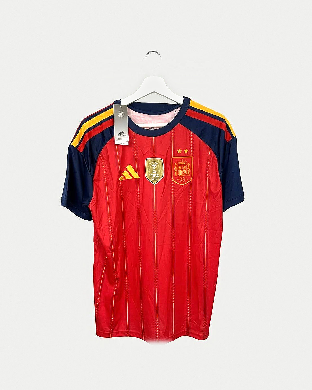 Camiseta de fútbol roja con rayas verticales a tono, mangas azul marino con franjas doradas en los hombros, cuello redondo azul marino, parche dorado de campeón del mundo y escudo con dos estrellas, colgada en una percha sobre pared clara
